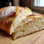 Close-up of a freshly baked, soft French bread loaf cut in half, showing the airy interior crumb and golden crust dusted with flour.