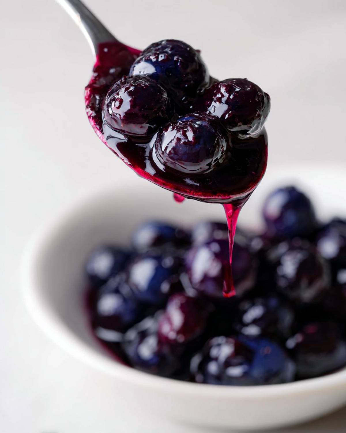 A metal spoon holds several plump blueberries coated in thick, glossy blueberry jam, dripping slightly over a white bowl of more berries.