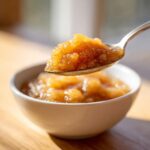 A spoonful of chunky, golden homemade applesauce being lifted from a white bowl.