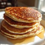 A tall stack of fluffy almond flour pancakes drizzled with maple syrup on a white plate.