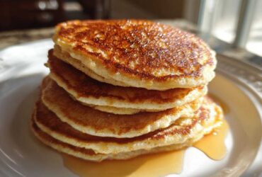 A tall stack of fluffy almond flour pancakes drizzled with maple syrup on a white plate.