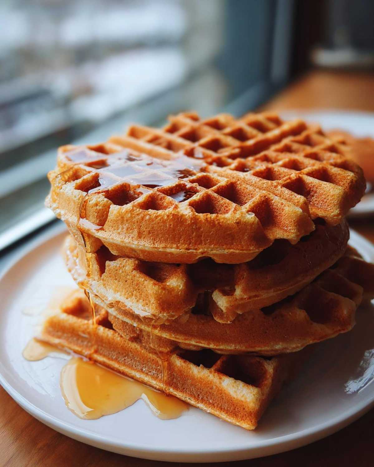 A tall stack of golden brown high protein waffles drizzled generously with maple syrup on a white plate.