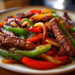 A close-up shot of sizzling steak fajitas featuring strips of glazed steak mixed with vibrant red, green, and yellow bell peppers.