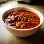 Close-up of a steaming white bowl filled with rich, thick crockpot chili featuring ground beef and kidney beans.