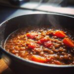 Close-up of a steaming bowl of hearty lentil soup with visible chunks of bright orange carrots and tomatoes.