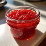 Close-up of a small glass jar filled with vibrant, chunky strawberry freezer jam, catching the sunlight.