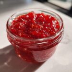 Close-up of a small glass jar filled with vibrant red strawberry freezer jam, catching bright sunlight.