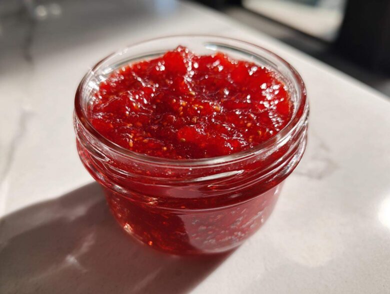 Close-up of a small glass jar filled with vibrant red strawberry freezer jam, catching bright sunlight.