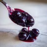 A spoonful of thick, glossy homemade blueberry jam dripping onto a light surface.