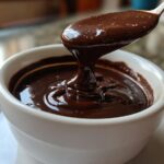 A spoonful of thick, glossy chocolate sauce recipe is being lifted from a white bowl, showing its rich texture.
