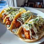 Close-up of three soft shell fish tacos filled with crispy fish, cabbage slaw, and drizzled with white sauce.