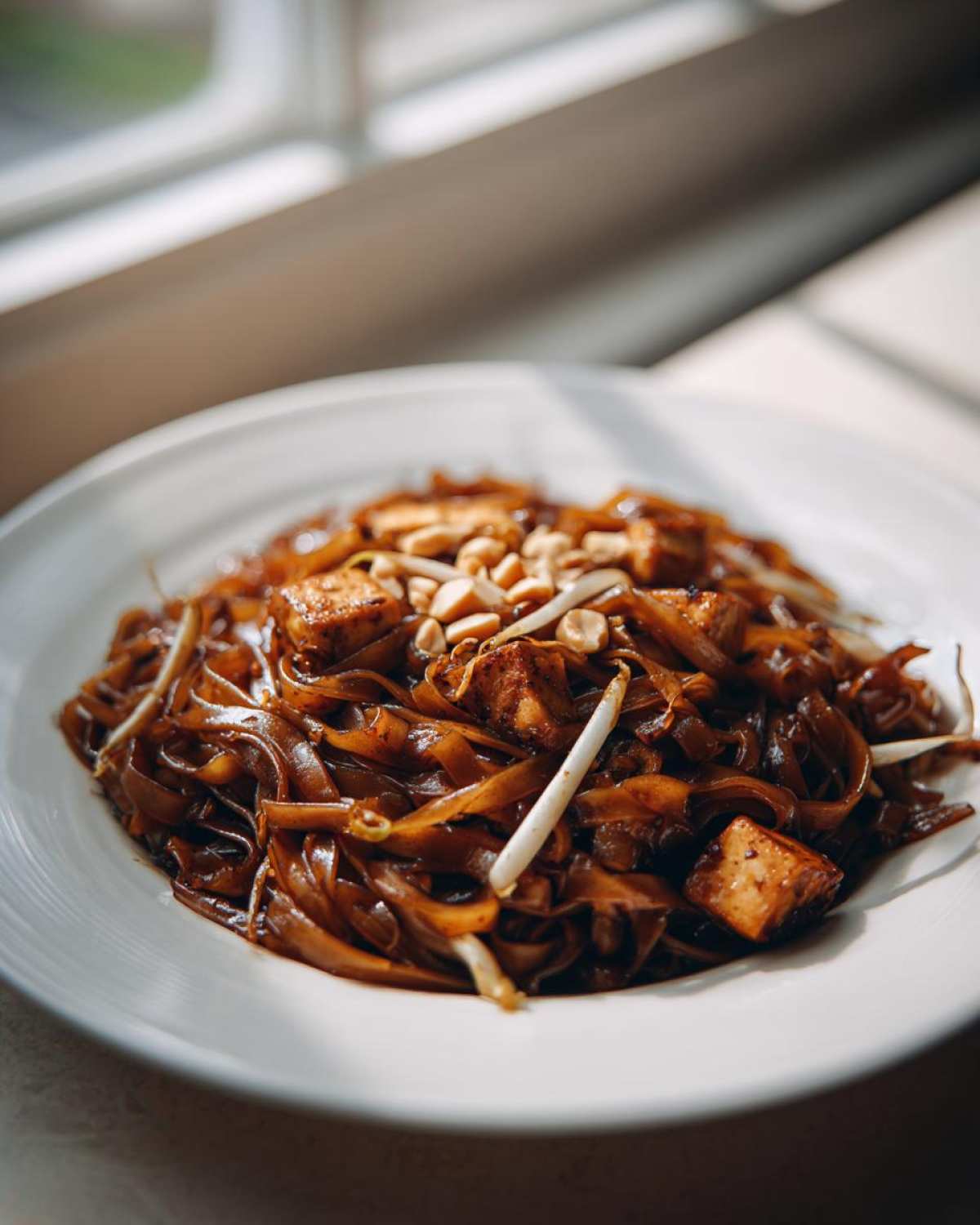 Close-up of rich, dark Pad Thai noodles with tofu cubes and peanuts, beautifully lit by natural light.