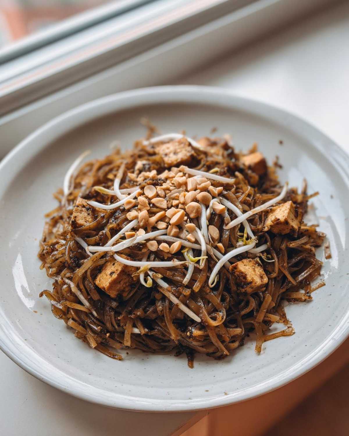 A close-up shot of a serving of dark, saucy Pad Thai noodles topped with crispy tofu cubes, fresh bean sprouts, and crushed peanuts.