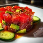 A vibrant, close-up shot of molded tuna tartare mixed with avocado chunks, garnished with sesame seeds and surrounded by cucumber slices.