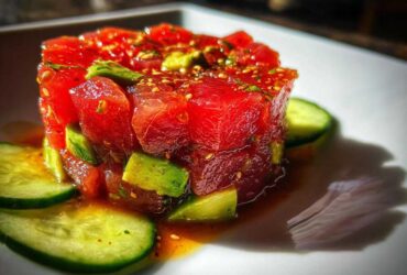 A vibrant, close-up shot of molded tuna tartare mixed with avocado chunks, garnished with sesame seeds and surrounded by cucumber slices.