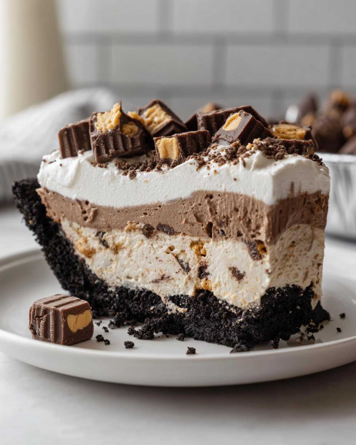 A tall slice of two-layer ice cream pie with a dark crust, topped with whipped cream and chopped peanut butter cups.