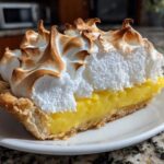 A close-up of a thick slice of lemon meringue pie showing bright yellow filling and tall, toasted meringue peaks.
