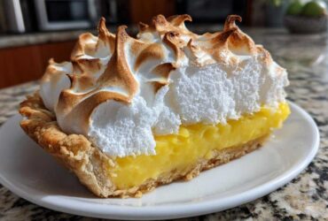 A close-up of a thick slice of lemon meringue pie showing bright yellow filling and tall, toasted meringue peaks.