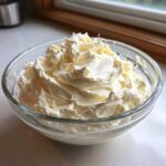 A clear glass bowl filled with freshly whipped, fluffy vanilla buttercream frosting.