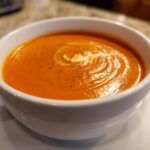 Close-up of a bowl of rich, orange tomato bisque, garnished with a swirl of cream and black pepper.