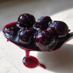 A silver spoon holds a dollop of thick, glossy blueberry jam with several whole blueberries visible, dripping onto a light surface. This is the amazing blueberry jam.