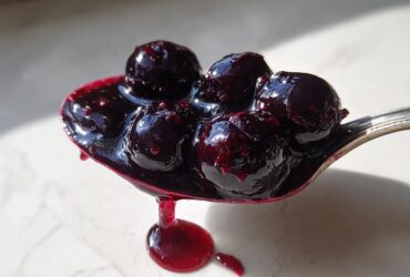 A silver spoon holds a dollop of thick, glossy blueberry jam with several whole blueberries visible, dripping onto a light surface. This is the amazing blueberry jam.