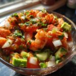 Close-up of a glass bowl filled with vibrant mexican shrimp cocktail, featuring shrimp, avocado, onion, and cilantro.