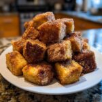 A mound of golden Air Fryer Crispy Cinnamon French Toast Bites heavily coated in cinnamon sugar, piled on a white plate.