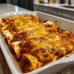 A close-up of freshly baked enchiladas covered in rich red sauce and melted cheddar cheese in a white baking dish.