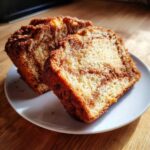 Two thick slices of moist Amish Cinnamon Bread with a visible cinnamon swirl and sugary crust, served on a white plate.