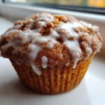 A single, freshly baked Apple Fritter Muffin topped with a thick, sweet white glaze and cinnamon sugar.