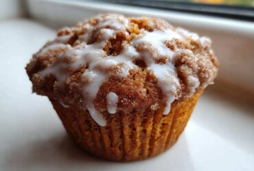 A single, freshly baked Apple Fritter Muffin topped with a thick, sweet white glaze and cinnamon sugar.