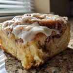 A close-up of a single, fluffy Banana Bread Batter Cinnamon Buns slice topped with thick white icing.