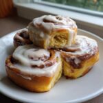 A stack of warm Banana Bread Cinnamon Rolls with Cream Cheese Frosting on a white plate.
