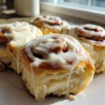 Close-up of four soft Banana Bread Cinnamon Rolls generously topped with dripping cream cheese frosting on a white plate.