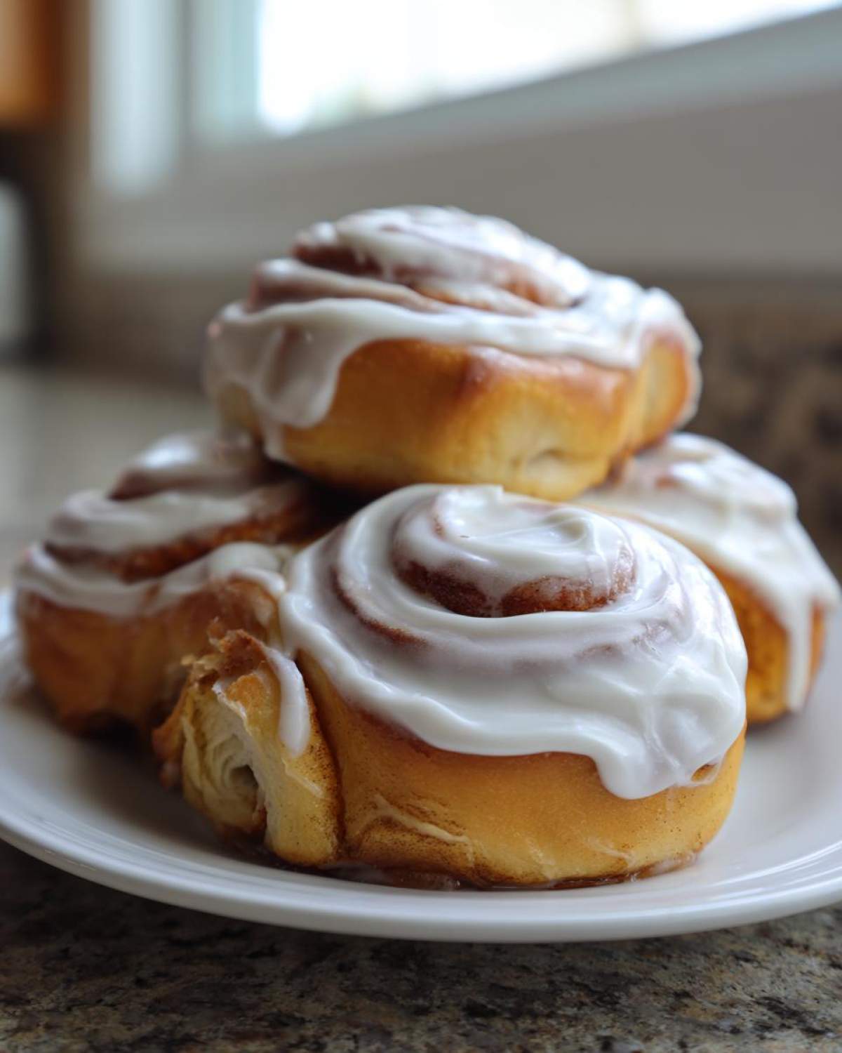 A stack of four freshly baked Banana Bread Cinnamon Rolls generously topped with white cream cheese frosting.