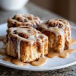 Three decadent Biscoff Cookie Butter Cinnamon Rolls generously drizzled with a thick, creamy glaze, sitting on a white plate.
