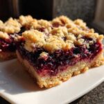Two squares of rich Blueberry Crumb Bars featuring a thick jammy filling and a golden buttery streusel topping, served on a white plate.