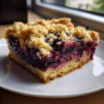 A square serving of Blueberry Crumb Bars with a thick, jammy berry filling and golden buttery streusel topping on a white plate.