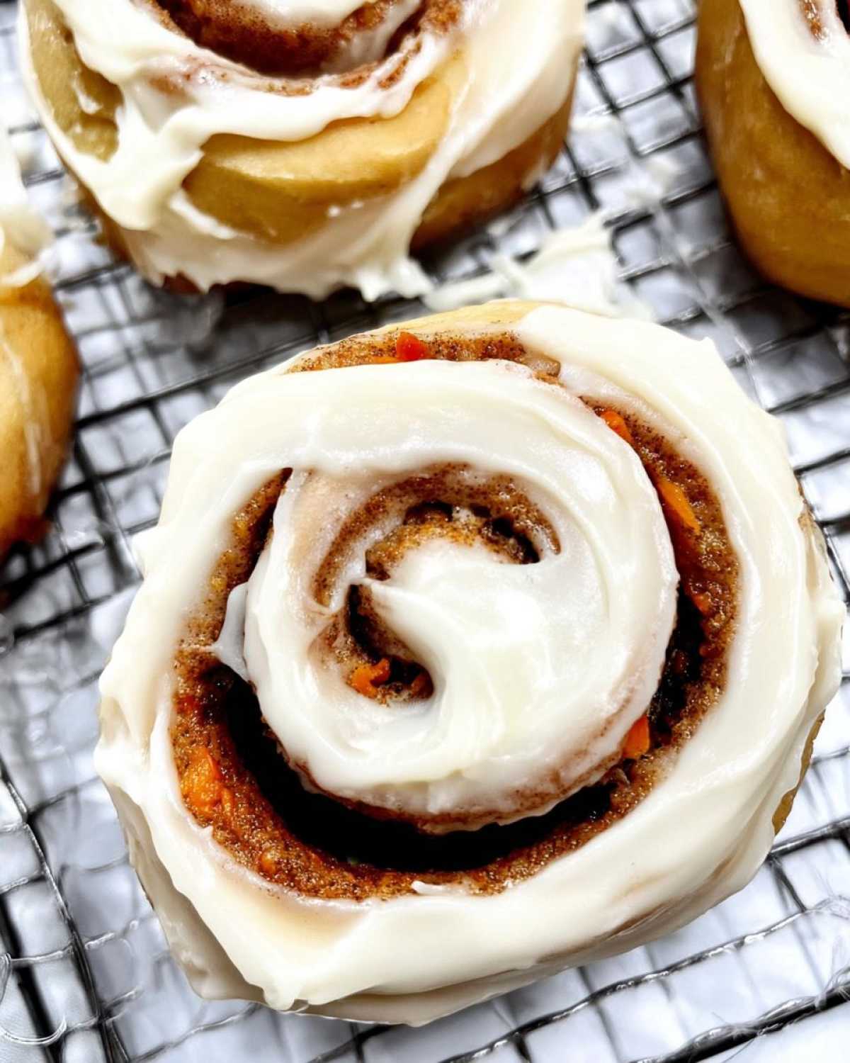A close-up of a freshly baked Carrot Cake Cinnamon Roll generously topped with creamy white frosting, resting on a cooling rack.