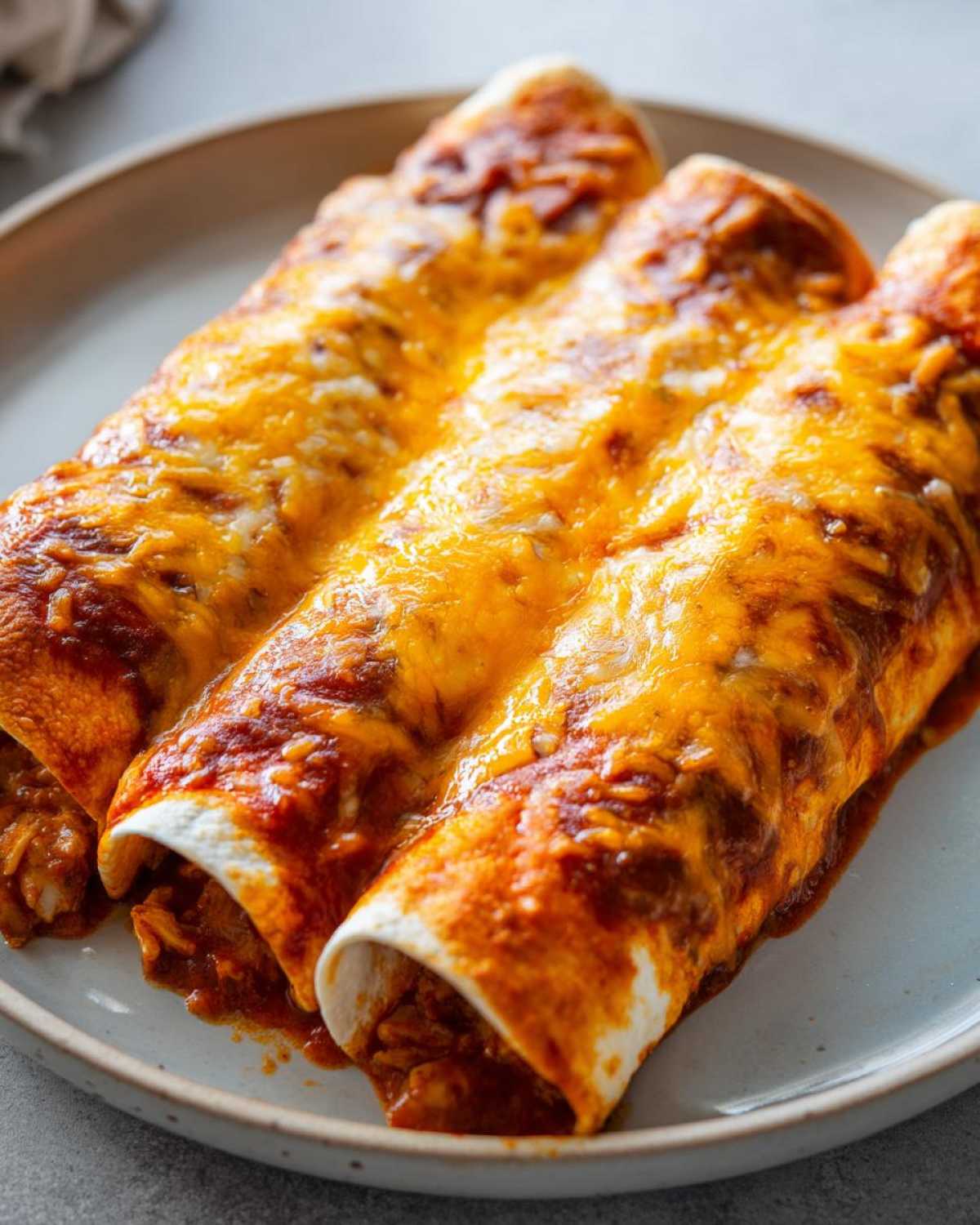 Close-up of three baked enchiladas smothered in red sauce and melted cheddar cheese, perfect for an Enchiladas Dinner.