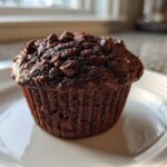 A single, dark Chocolate Banana Muffin topped with melted chocolate chips sitting on a white plate.