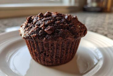 A single, dark Chocolate Banana Muffin topped with melted chocolate chips sitting on a white plate.