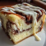 A close-up of a rich slice of cinnamon roll cheesecake dessert drizzled with white icing.