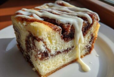 A close-up of a rich slice of cinnamon roll cheesecake dessert drizzled with white icing.