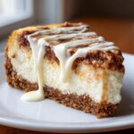 A decadent slice of cinnamon roll cheesecake featuring a thick graham cracker crust, creamy filling, cinnamon swirl, and white icing drizzle.