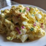 Close-up of Classic Macaroni Salad Creamy Summer Side, featuring elbow macaroni coated in yellow dressing with chunks of egg and red onion.