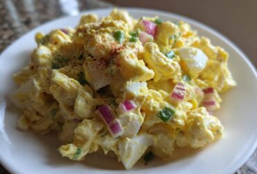 Close-up of Classic Macaroni Salad Creamy Summer Side, featuring elbow macaroni coated in yellow dressing with chunks of egg and red onion.