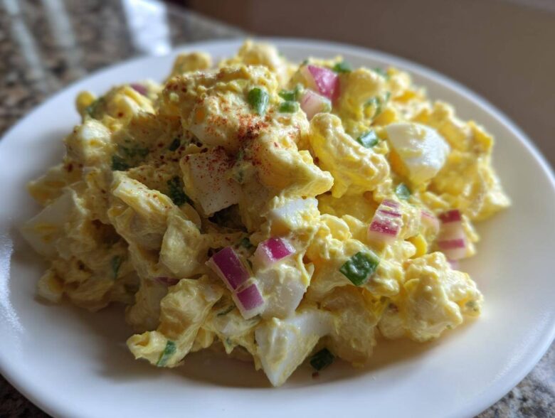 Close-up of Classic Macaroni Salad Creamy Summer Side, featuring elbow macaroni coated in yellow dressing with chunks of egg and red onion.