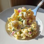 A fork lifts a perfect bite of Classic Macaroni Salad Creamy Summer Side, showing elbow macaroni, egg, and red onion.